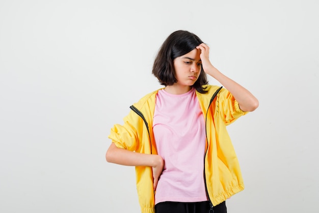 Menina adolescente em t-shirt, jaqueta coçando a cabeça e olhando pensativa, vista frontal.