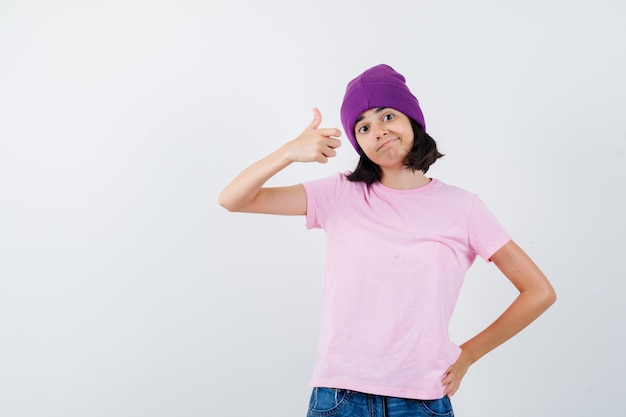 Menina adolescente em t-shirt rosa, gorro, jeans aparecendo o polegar, segurando a mão na cintura e olhando alegre, vista frontal.