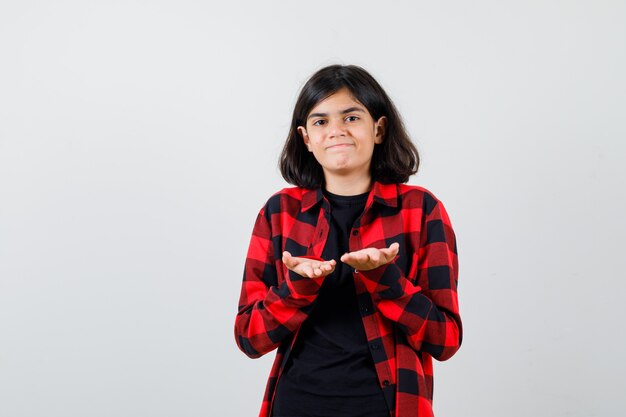 Menina adolescente fazendo gesto de dar ou receber em t-shirt, camisa quadriculada e parecendo satisfeito, vista frontal.