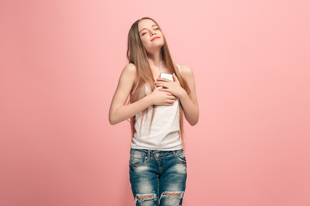 Menina adolescente feliz em pé, sorrindo com o celular sobre o fundo rosa da moda do estúdio. Belo retrato feminino de meio corpo. Emoções humanas, conceito de expressão facial.