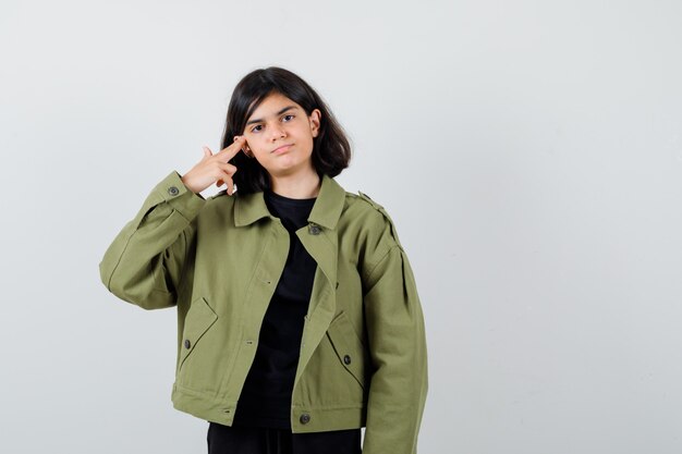 Menina adolescente mostrando gesto de arma em t-shirt, jaqueta verde e parecendo autoconfiante, vista frontal.