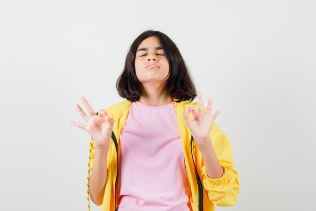 Menina adolescente mostrando o gesto ok com os olhos fechados em agasalho amarelo, camiseta e parecendo cansado, vista frontal.