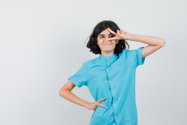 Menina adolescente mostrando o sinal-V no olho em uma camisa azul e olhando feliz.