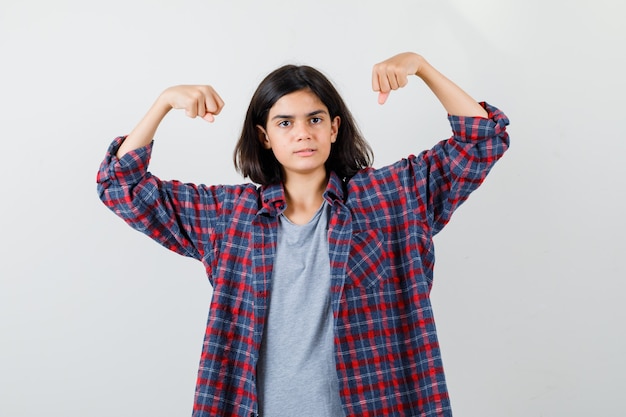 Menina adolescente mostrando os músculos dos braços em roupas casuais e olhando forte, vista frontal.