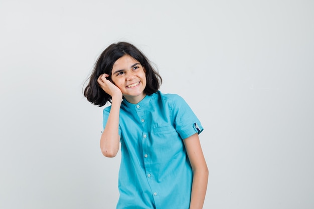 Menina adolescente segurando a mão em sua bochecha enquanto sorri de camisa azul e parece feliz.