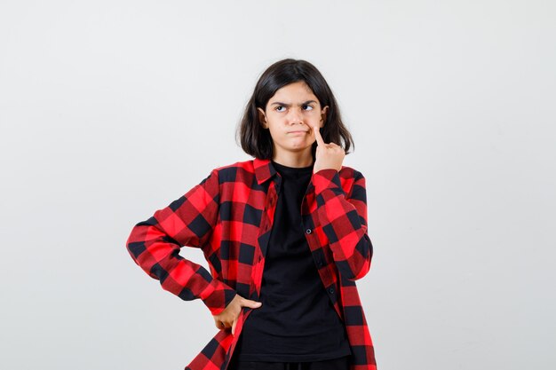 Menina adolescente segurando o dedo mindinho na bochecha em t-shirt, camisa quadriculada e parecendo insatisfeito, vista frontal.