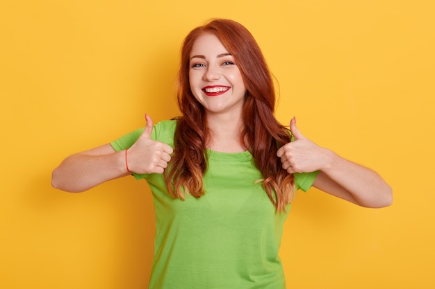 Menina adorável alegre mostrando o polegar para cima enquanto posa isolada, ruiva, com uma camiseta verde