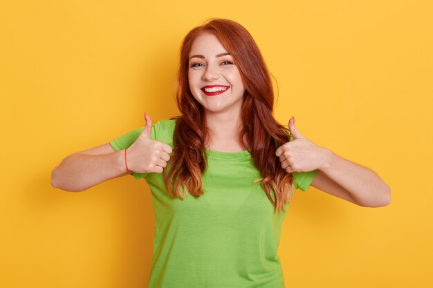 Menina adorável alegre mostrando o polegar para cima enquanto posa isolada, ruiva, com uma camiseta verde