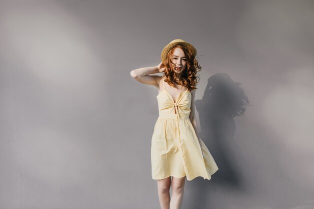 Menina adorável magro com cabelo encaracolado, posando em um vestido amarelo. Retrato do modelo feminino caucasiano com chapéu de palha isolado na parede cinza.