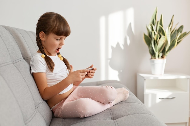 Menina adorável surpresa com rabo de cavalo sentado no sofá na luz da sala de estar com telefone inteligente nas mãos, olhando para a tela do telefone móvel com a boca aberta, expressando espanto.