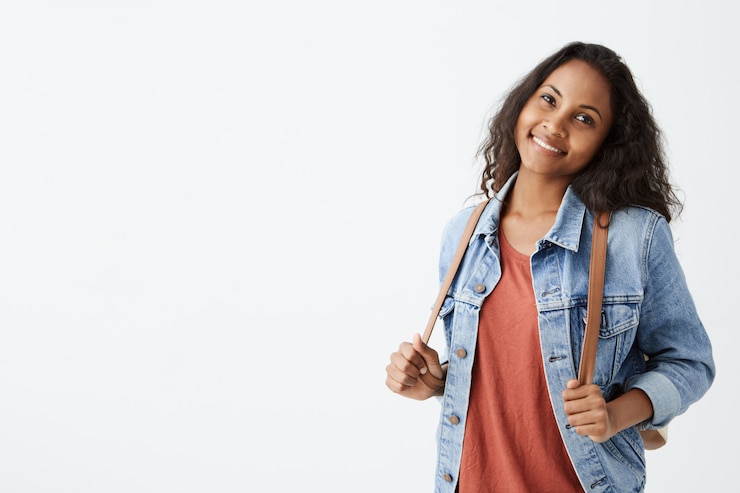 Foto grátis menina afro-americana elegante com cabelos escuros ondulados se divertindo dentro de casa e com um sorriso alegre. jovem bonita jaqueta jeans sorrindo alegremente mantendo as mãos na mochila