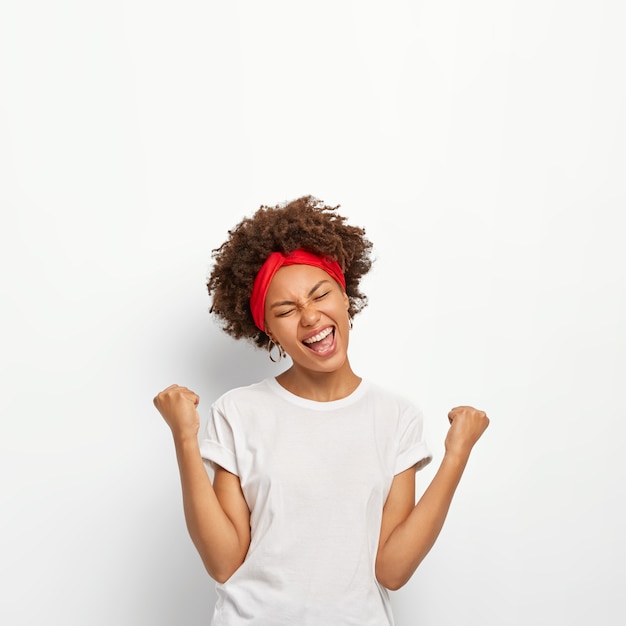 Menina Afro feliz e encantada fecha os punhos, sente triunfo, alegra-se com a vitória, mantém os olhos fechados, sorri amplamente, veste uma camiseta branca, fica em pé dentro de casa