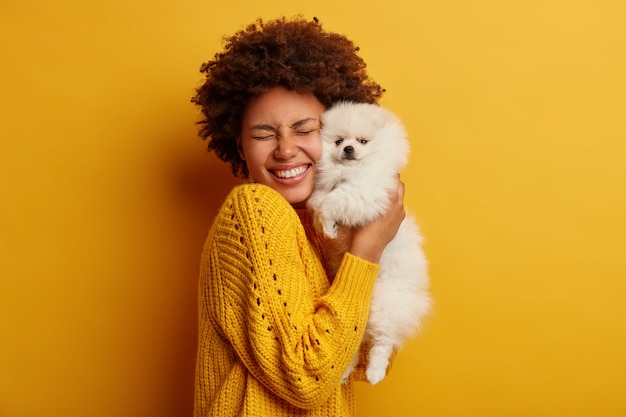 Foto grátis menina afro feliz e satisfeita ganha um cachorrinho adorável, brinca e abraça com amor o amigo de quatro patas, em pé contra um fundo amarelo