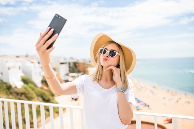 Menina agradável de óculos escuros e chapéu tocando sua bochecha enquanto faz uma selfie no mar