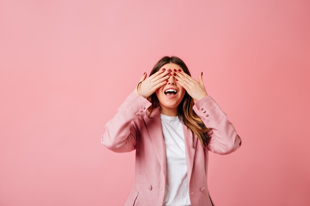 Menina alegre, cobrindo os olhos com as mãos. Foto de estúdio de mulher elegante casaco isolada no fundo rosa.