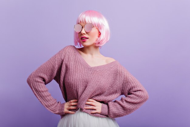 Menina alegre com cabelo reto rosa em pé em pose confiante e sorrindo. senhora muito europeia de suéter e óculos brilhantes, dançando durante a sessão de fotos.