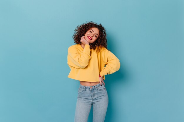 Menina alegre com lábios vermelhos e sorriso branco como a neve Olha para a câmera no espaço azul. Retrato de uma senhora encaracolada bonita no top amarelo com jeans e mangas compridas.