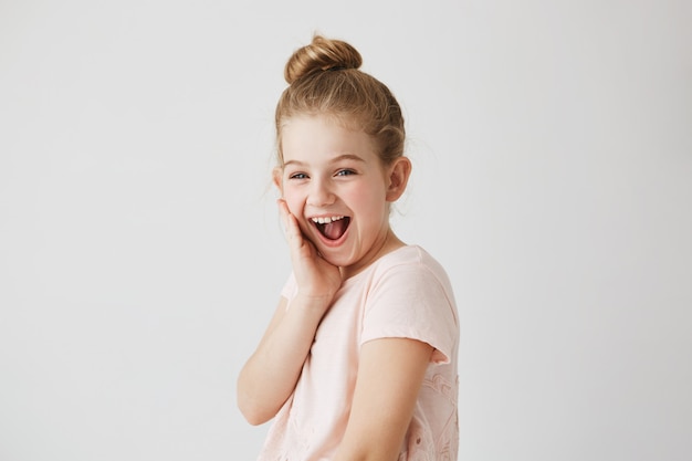 Menina alegre com olhos azuis e penteado coque com expressão feliz depois de se ver no espelho com um belo penteado novo.