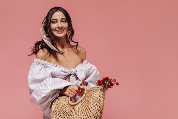 Foto grátis menina alegre com penteado encaracolado moderno em blusa lilás de manga larga segurando saco com flores vermelhas e olhando para a câmera