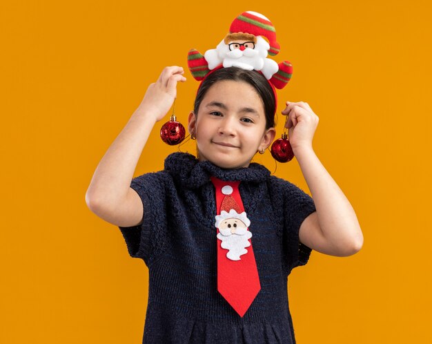 Menina alegre com vestido de tricô usando gravata vermelha com aro engraçado na cabeça segurando bolas de natal nas orelhas sorrindo em pé sobre a parede laranja