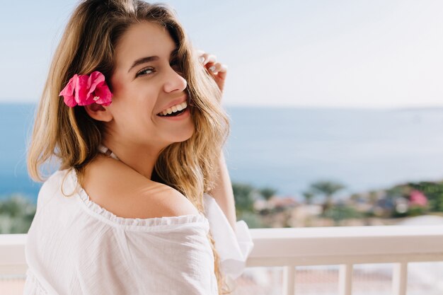 Menina alegre da risada com uma flor bonita no cabelo castanho claro, posando com vista para o horizonte. Linda jovem vestida de branco aproveitando as férias no resort e tomando ar fresco