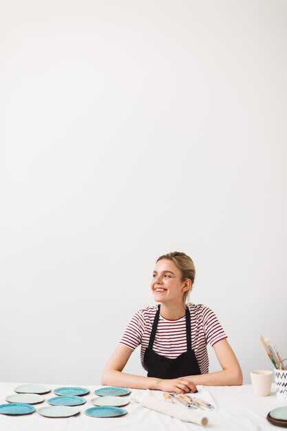 Menina alegre de avental preto e camiseta listrada, sentado à mesa com pratos artesanais alegremente olhando de lado no estúdio de cerâmica