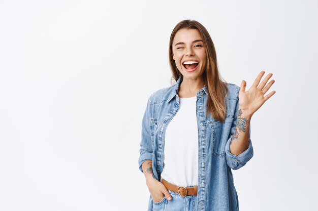 Menina alegre e amigável dizendo olá, piscando e sorrindo, acenando com a mão na frente, dizer olá e cumprimentar você, encostada na parede branca