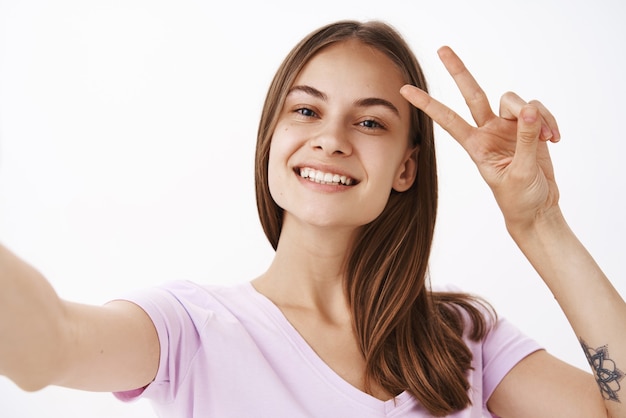 Menina alegre e charmosa, de aparência amigável, com cabelo castanho e um sorriso largo, mostrando o sinal de paz ou vitória perto do rosto, enquanto tira uma selfie com o smartphone na parede cinza