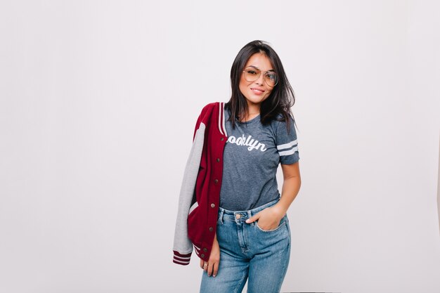 Menina alegre e magro em camiseta cinza e calça jeans posando com a mão no bolso e sorrindo. Modelo feminino de cabelos negros em jeans e homem-bomba em pé.