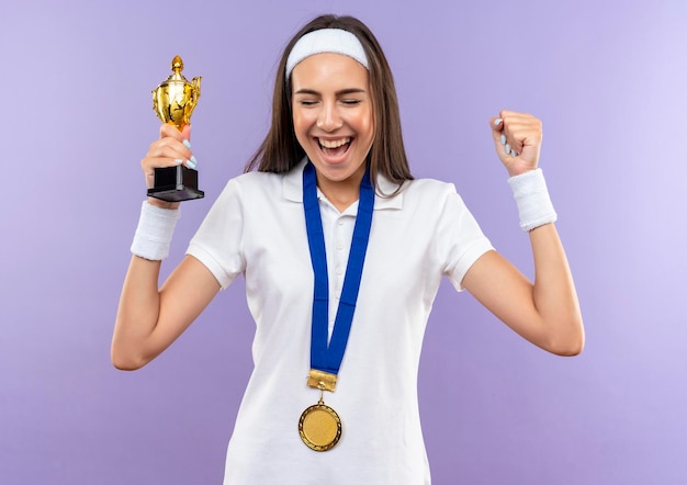 Foto grátis menina alegre e muito esportiva usando bandana, pulseira e medalha, segurando o copo levantando o punho com os olhos fechados, isolada na parede roxa