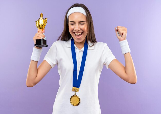 Menina alegre e muito esportiva usando bandana, pulseira e medalha, segurando o copo levantando o punho com os olhos fechados, isolada na parede roxa