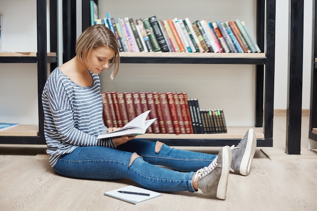 Menina alegre jovem estudante de cabelos claros, com cabelos curtos em camisa listrada e calça jeans, sentada no chão na biblioteca, lendo o livro, gastando tempo produtivo após o estudo, se preparando para os exames.