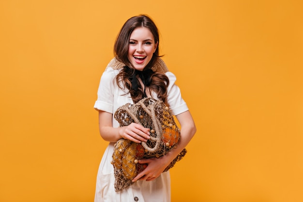 Menina alegre posando com uma sacola cheia de frutas. mulher de vestido branco posando em fundo laranja.