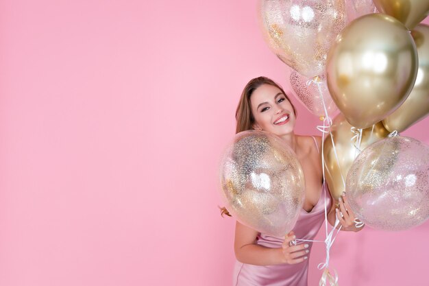 Menina alegre segurando muitos balões de ar dourados na festa de aniversário de ano novo com fundo rosa Foto Premium