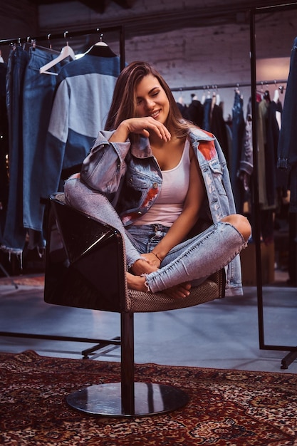 Menina alegre vestindo um jeans angustiado e jaqueta jeans, sentado em uma cadeira no provador de uma loja de roupas.