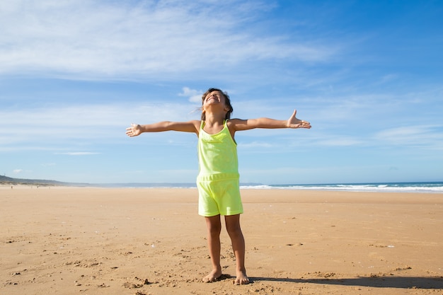 Foto grátis menina animada com roupas de verão, em pé com as mãos voadoras na praia e virando o rosto para cima