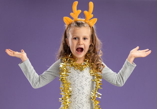 Menina animada com uma argola de cabelo de natal e uma guirlanda no pescoço estendendo as mãos isoladas sobre fundo azul