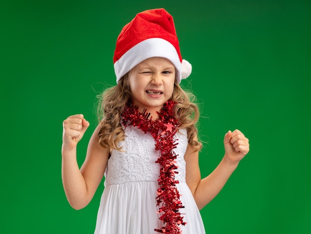 Menina animada e piscando usando chapéu de Natal com guirlanda no pescoço mostrando gesto de sim isolado na parede verde