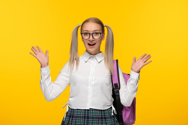 Menina animada loira do dia mundial do livro com mochila em papel de parede amarelo com óculos