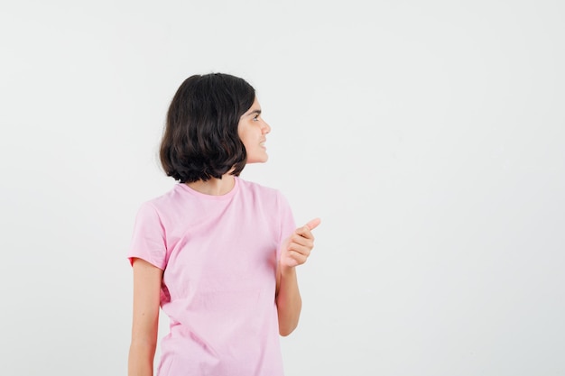 Menina aparecendo o polegar, olhando de lado em uma camiseta rosa e olhando otimista, vista frontal.