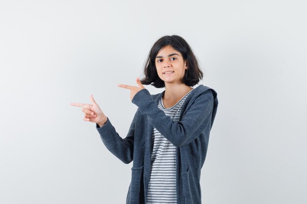 Menina apontando de lado em t-shirt, jaqueta e olhando alegre.