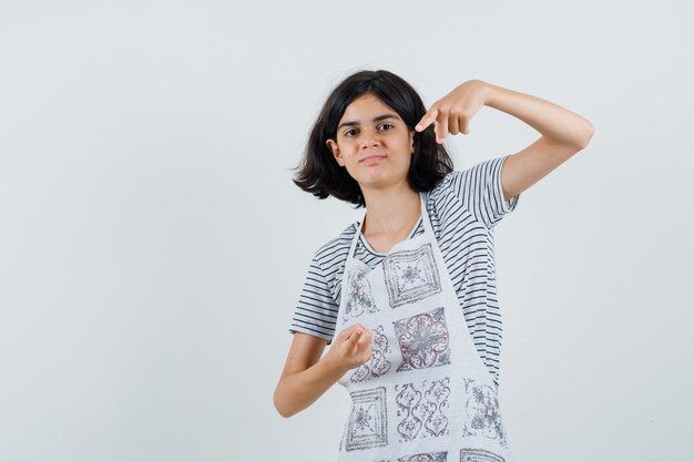 Menina apontando para a câmera em t-shirt, avental e parece confiante.