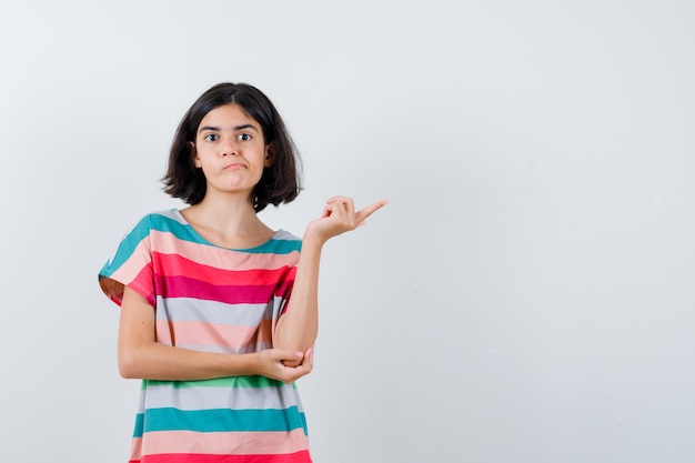 Menina apontando para a direita, segurando a mão no cotovelo em t-shirt, jeans e olhando séria, vista frontal.