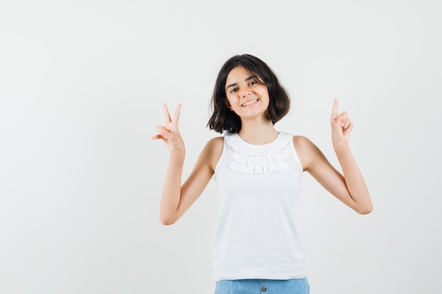 Menina apontando para cima, mostrando o sinal v de blusa branca, shorts e parecendo alegre. vista frontal.