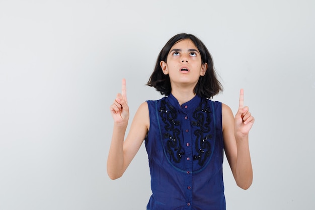 Menina apontando para cima na blusa azul e olhando com foco.