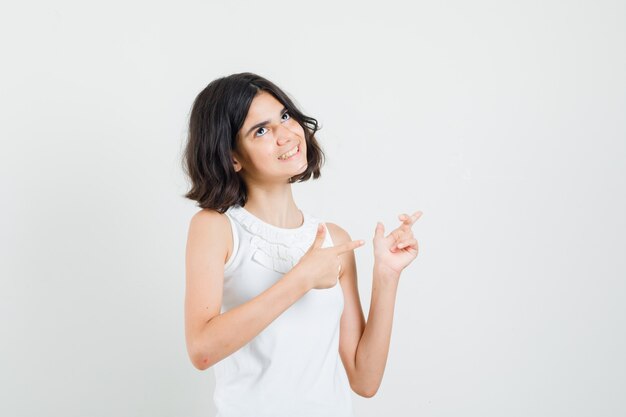 Menina apontando para o canto superior direito na blusa branca e olhando alegre, vista frontal.