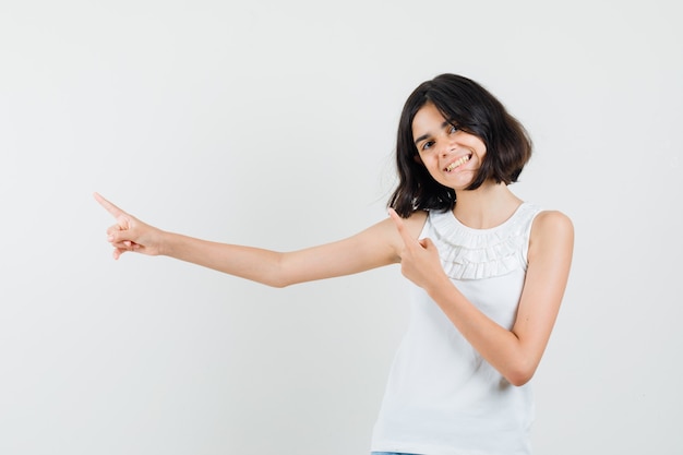 Menina apontando para o canto superior esquerdo na blusa branca e olhando alegre, vista frontal.