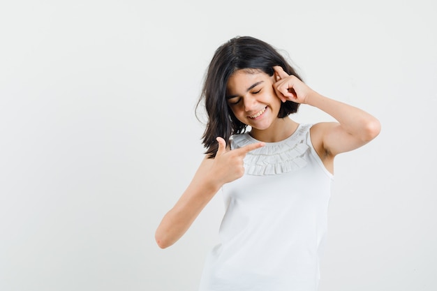 Menina apontando para o lado na blusa branca e parecendo alegre. vista frontal.