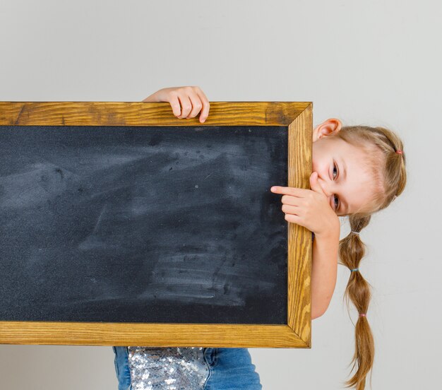 Menina, apontando para o quadro-negro em t-shirt e jeans saia e olhando feliz, vista frontal.