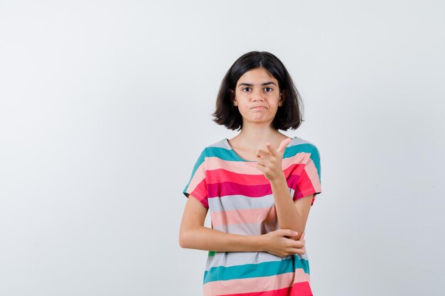 Menina apontando, segurando a mão no cotovelo em t-shirt, jeans e parecendo descontente, vista frontal.